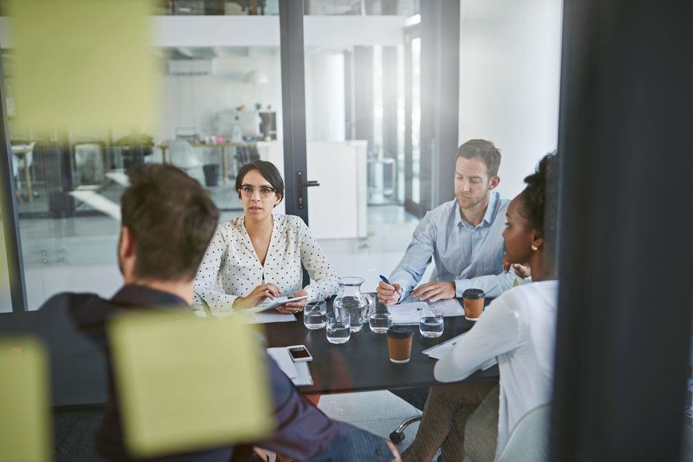 A group of engineers in a conference room conducting a design review meeting, discussing documents and taking notes around a table with laptops, papers, and coffee cups.