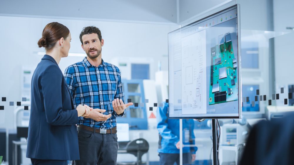 Female Computer Engineer and Male Project Manager Use Digital Interactive Whiteboard That Shows 3D Printed Circuit Board Concept. Developers Find Problem Solution. Modern Factory Meeting Room